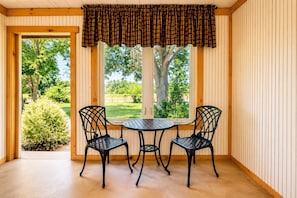 Una habitación con un revestimiento de madera, una ventana con vistas a follaje, y dos sillas de metal negro alrededor de una pequeña mesa redonda.