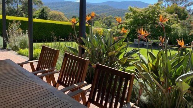 A covered outdoor seating area with wooden chairs and a table, overlooking a landscaped garden with various plants and a distant mountain range. - Mon Desir - Yarra Valley (Yarra Ranges)