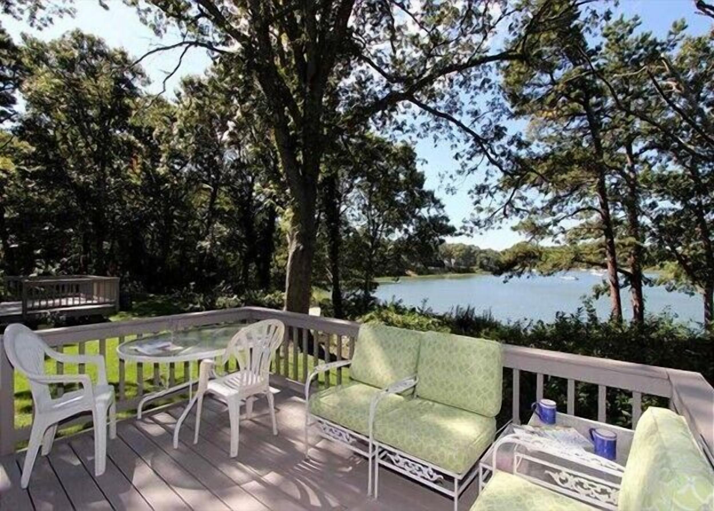 A deck with white chairs and a table, overlooking a lake surrounded by trees.