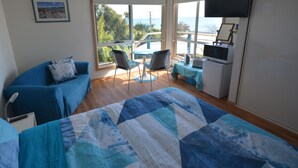 Ein Zimmer mit einem Bett, einem blauen Sofa, einem Esstisch mit Stühlen, einem Fernseher und Blick auf das Meer.