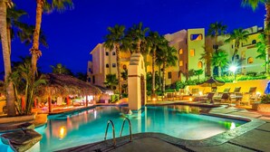 A nighttime resort pool area with a clear pool, lounge chairs, palm trees, and a building with multiple balconies. - Casa Mar Azul, a Stunning Oceanfront Condo (Cabo San Lucas)