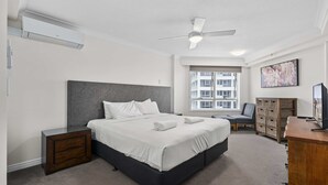 Ein modernes Schlafzimmer mit einem großen Bett, einem Deckenventilator, einer Klimaanlage, einem Nachttisch aus Holz, einem Schreibtisch mit Stuhl, einem Fernseher und einem Fenster mit Blick auf Gebäude.