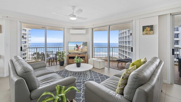 Ein modernes Wohnzimmer mit einer grauen Sofagarnitur, einem Couchtisch und einem Fernseher. Das Zimmer verfügt über große Fenster mit Blick auf den Strand und einen Balkon mit Sitzgelegenheit.