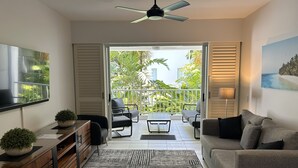 Un salon moderne avec un canapé, une table basse et un téléviseur posé sur un meuble en bois. Il y a un balcon avec vue sur la verdure et un ventilateur de plafond.