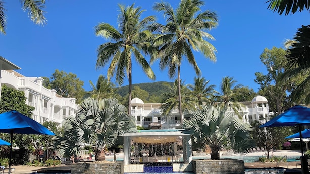 Un espace piscine doté de chaises longues et de parasols, entouré de palmiers et bordé d’un bâtiment blanc surmonté d’une coupole.