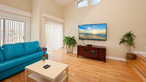 Una sala de estar con un sofá azul, una mesa de centro, un televisor montado en la pared y dos plantas en macetas.