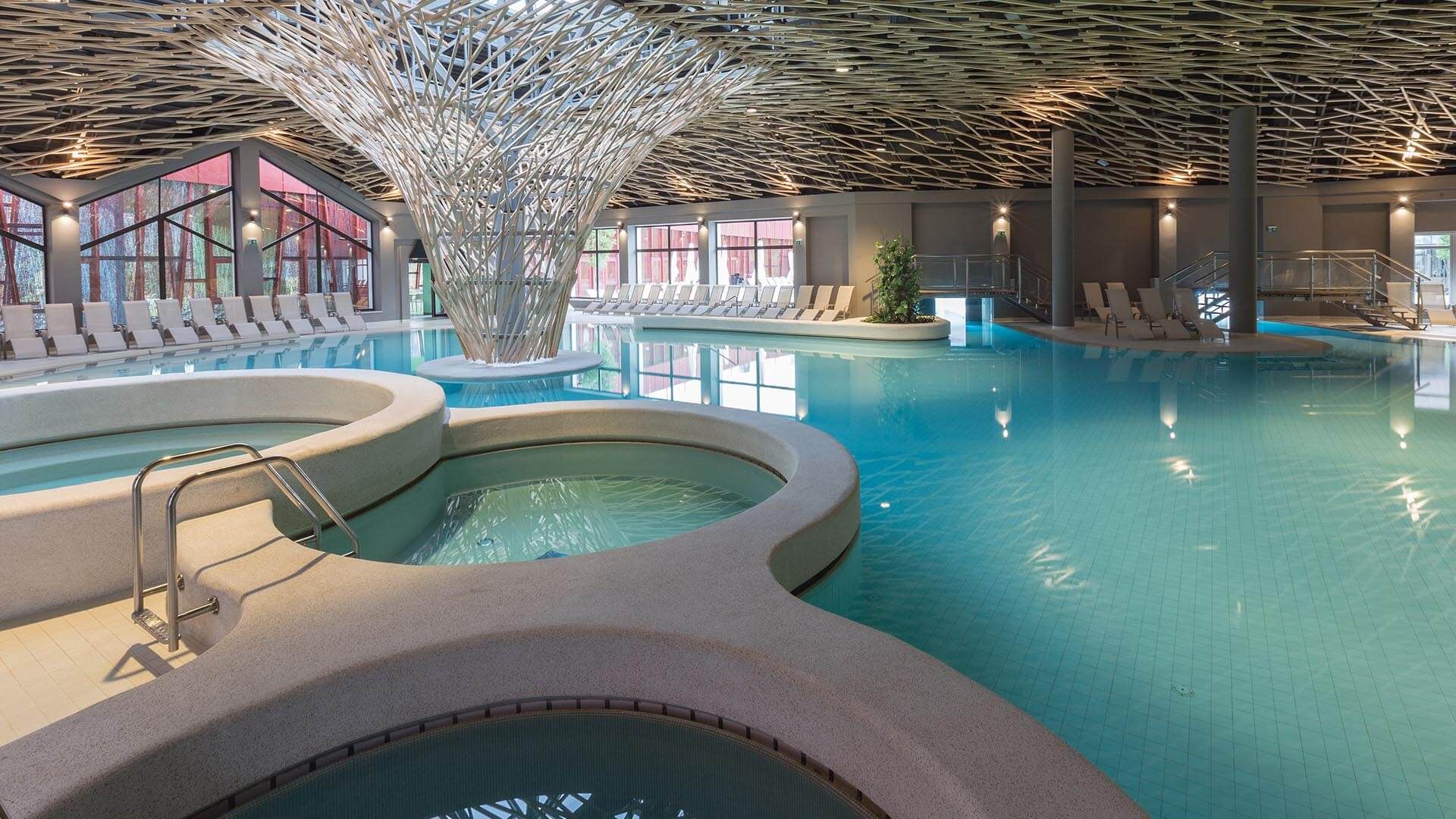 Una piscina interior con múltiples carriles, un elemento acuático central y zonas de descanso con asientos.