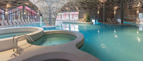 Una piscina interior con múltiples carriles, un elemento acuático central y zonas de descanso con asientos.