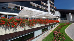 Ein modernes Hotel mit mehreren Balkonen, geschmückt mit roten Blumen und weißen Sonnenschirmen, und einem gepflegten Rasen.