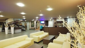 Un vestíbul d'hotel amb zones d'estar, una taquilla de recepció, una escala i una pantalla de televisió.