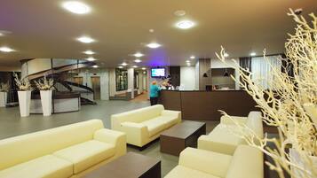 A hotel lobby with seating areas, a reception desk, a staircase, and a television screen.