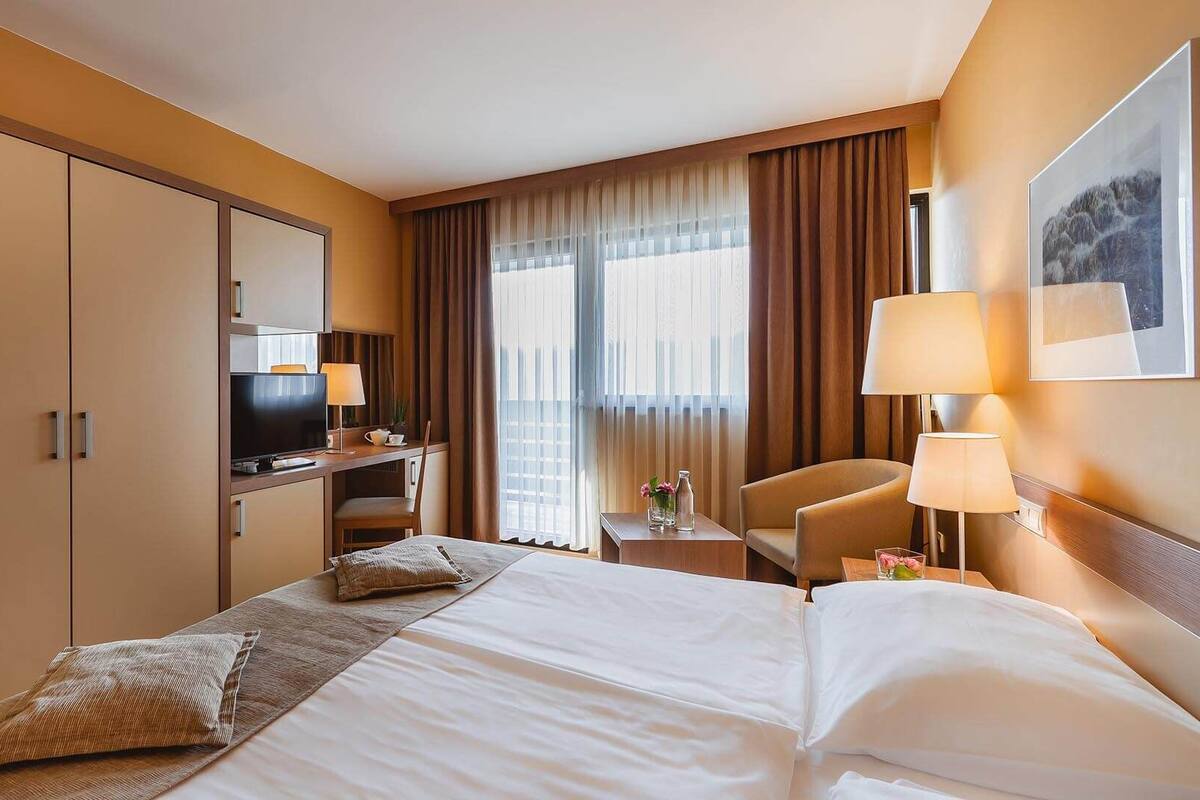 Habitación de hotel con una cama grande, un escritorio con televisor, una silla y una ventana con cortinas.
