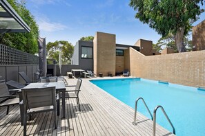 Ein Schwimmbecken mit Holzterrasse, Liegestühlen und einem modernen Gebäude mit Backsteinfassade.