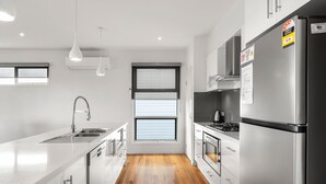 Eine moderne Küche mit einem Edelstahl-Kühlschrank, einem Einbauofen und einem Gasherd. Ein Fenster mit Jalousien, eine Spüle mit Armatur und Pendelleuchten sind vorhanden.
