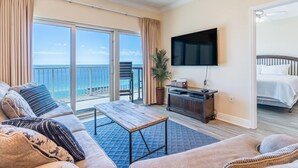 A spacious living room with a large sofa, a coffee table, and a flat-screen TV. There is a balcony with a view of the ocean.