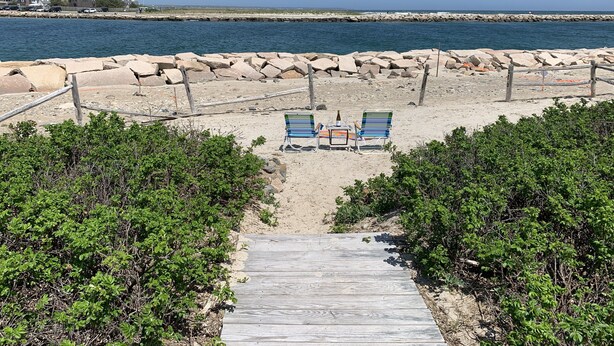 Een strand met twee blauwe ligstoelen en een fles op een klein tafeltje, een houten pad dat naar het strand leidt en een rotsachtige zeewering op de achtergrond.
