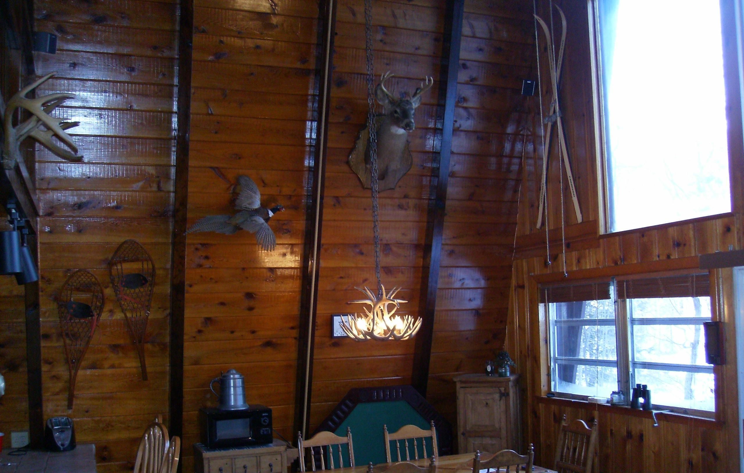 A rustic cabin interior with wooden walls, a dining table, chairs, a small kitchen area with a microwave, and a window.