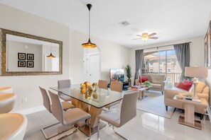 A modern living room with a glass dining table, comfortable seating, a ceiling fan, and a large window with curtains.