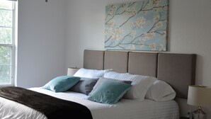 Una cama bien tendida con un plaid oscuro, una mesita de noche con una lámpara y un cuadro de flores de cerezo en la pared.