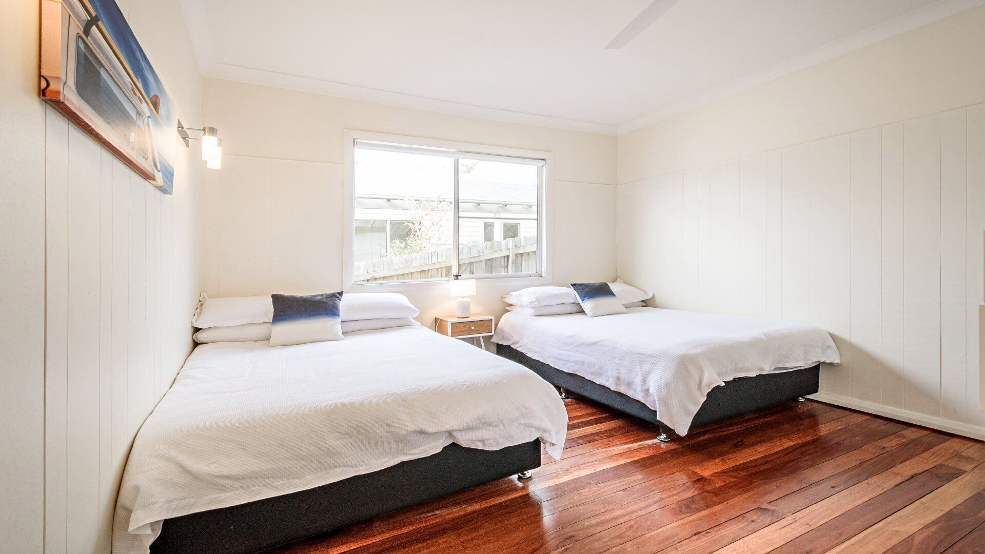 A bedroom with two beds, a window, a nightstand, and a wall-mounted lamp.
