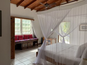 A bedroom with a bed, a window with curtains, a bench with red cushions, and a ceiling fan.