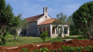 Um edifício rústico de pedra com telhado de telhas vermelhas e janelas em arco, rodeado por vegetação e uma área cercada.