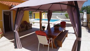 Um pátio externo com uma mesa de madeira, cadeiras brancas, um grande guarda-sol e uma piscina ao fundo.