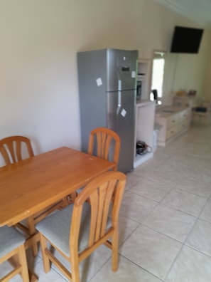 A dining area with a wooden table and chairs, a grey refrigerator, and a kitchen area with cabinets and a TV mounted on the wall.