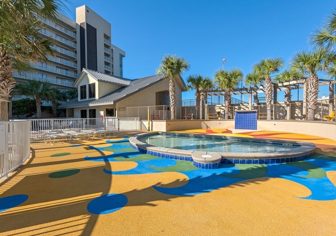 A swimming pool with colorful play area tiles, a building with balconies, and palm trees.