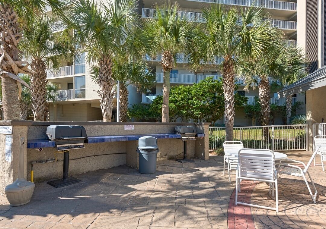 An outdoor patio with a barbecue grill, seating area, and palm trees.