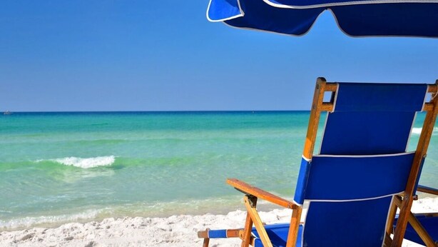 Ein Liegestuhl und Sonnenschirm an einem sandigen Strand mit klarem türkisblauem Wasser und wolkenlosem Himmel.
