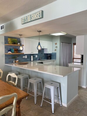 Una cocina moderna con una isla central, taburetes blancos y un cartel que dice 'LET'S DO SUNSET'.