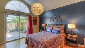 Quarto com cama, duas mesas de cabeceira, ventilador de teto e vista para uma palmeira através de uma porta de vidro deslizante.