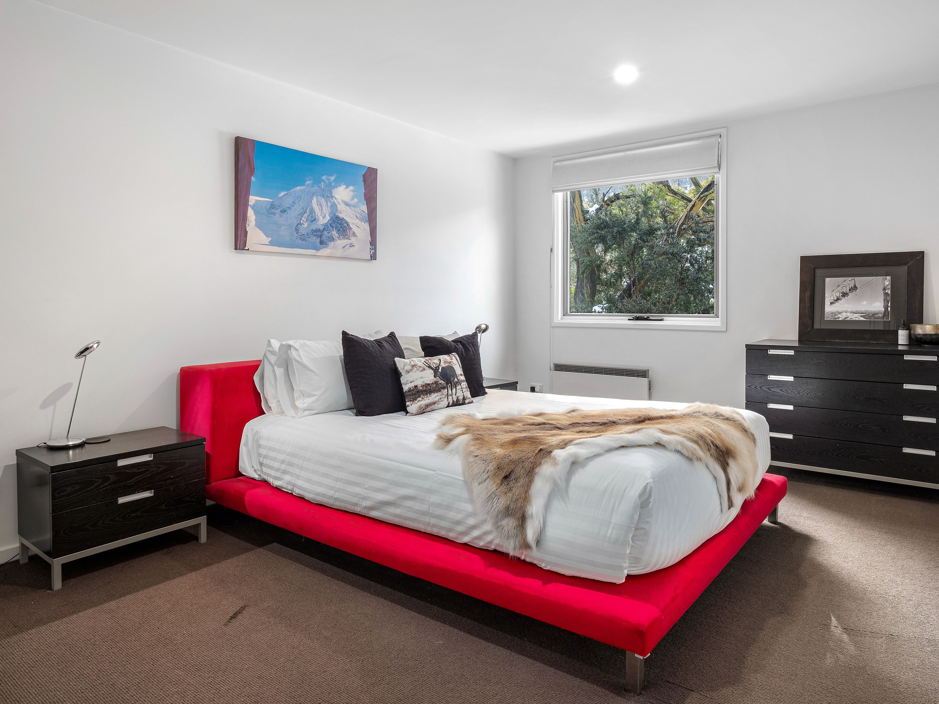 A modern bedroom with a red bed, white bedding, a mounted picture, a window with a view of trees, and a dark wooden dresser.