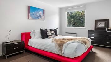 A modern bedroom with a red bed, white bedding, a mounted picture, a window with a view of trees, and a dark wooden dresser.