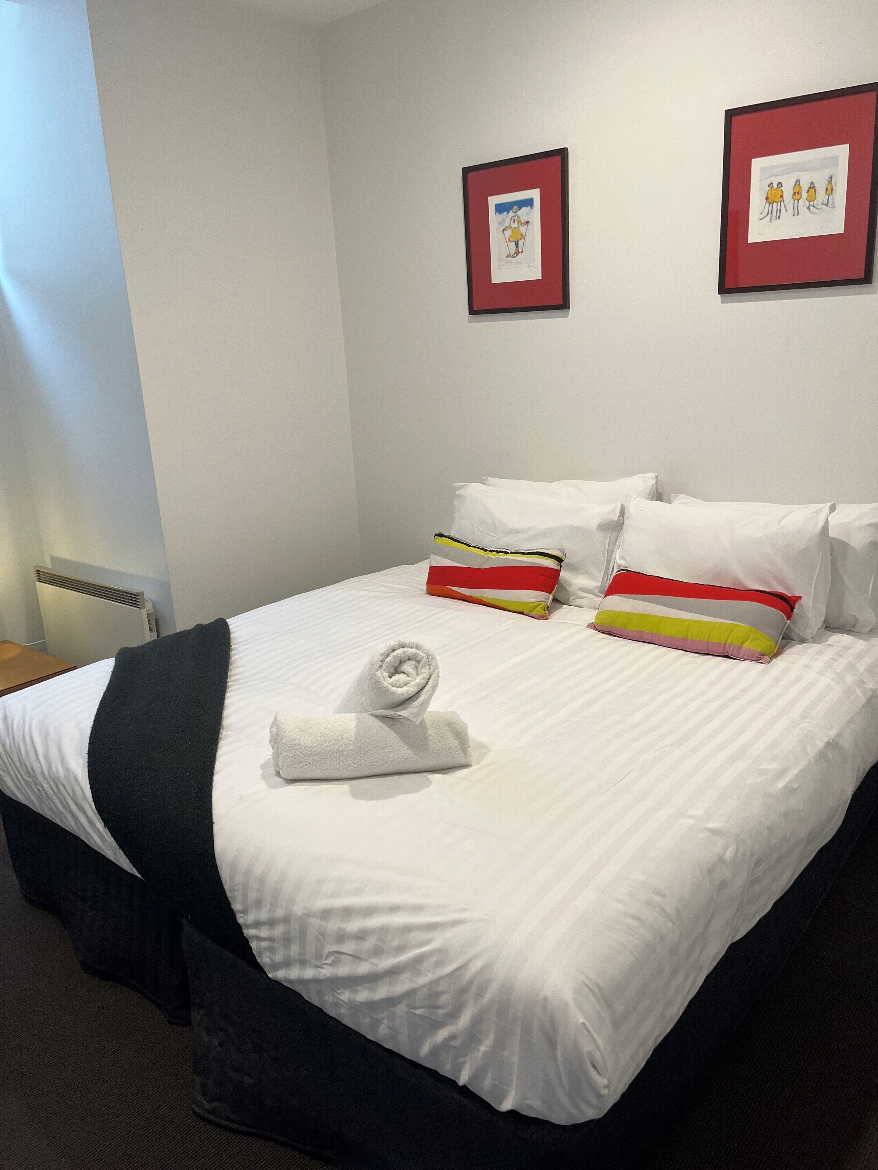 A neatly made bed with white linens, a rolled towel, and colorful pillows. There are two framed artworks on the wall.