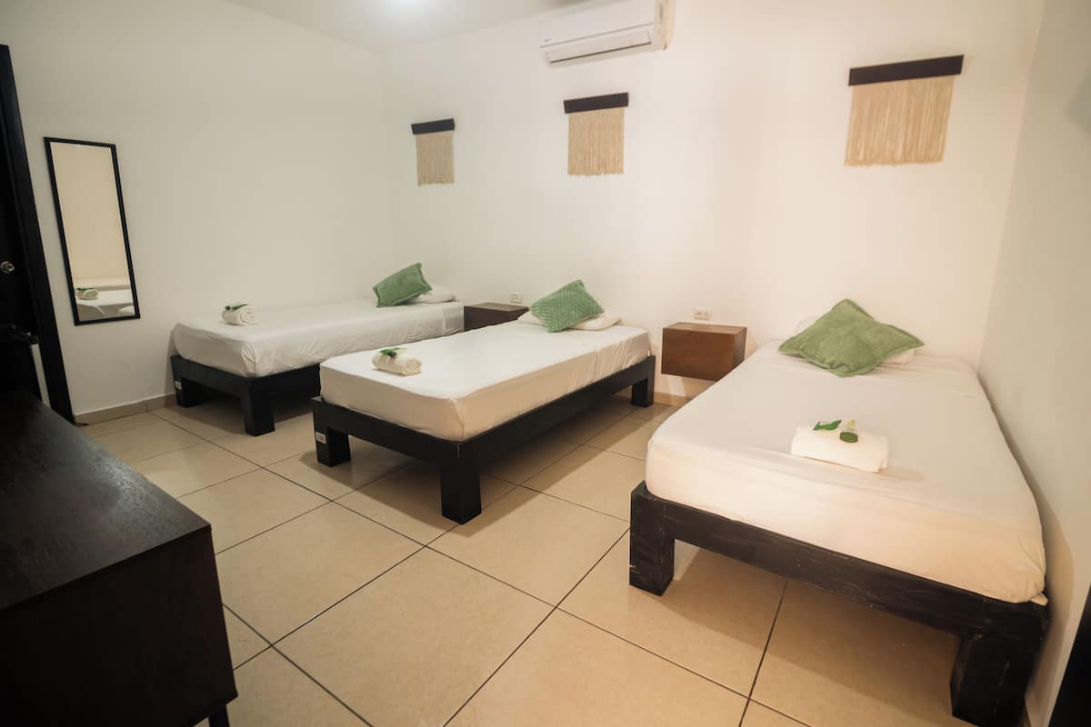 Ein Hotelzimmer mit drei Einzelbetten, einem Holzkoffer und einem großen Spiegel.