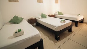 A room with three single beds, each with a white mattress and green pillow. There are wooden nightstands with a small table between the beds. The room has a tiled floor and plain white walls.