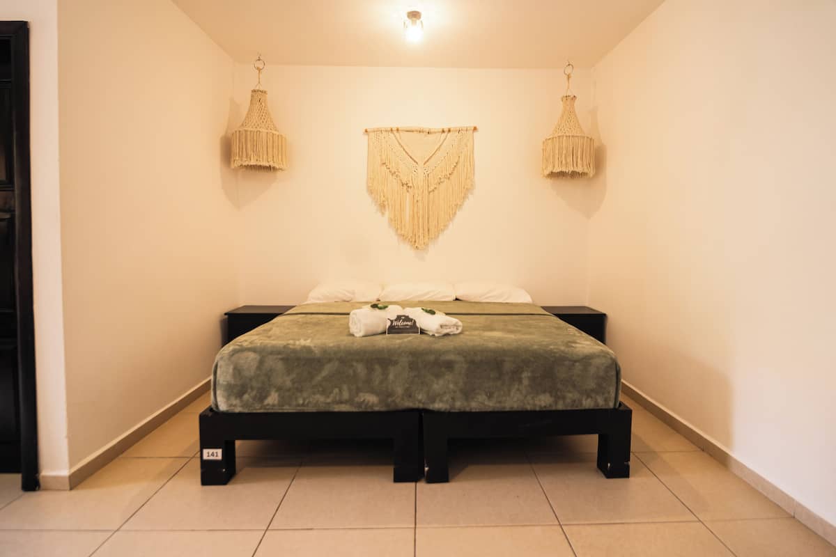 Ein ordentlich bezogenes Bett mit einer grünen Tagesdecke, zwei Nachttischen und einem Wandbild aus Makramee darüber.