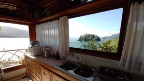 A kitchen with wooden cabinets, a double sink, and a view of a lake and mountains through a large window. - The most beautiful view in Ilhabela (IlhaBela)