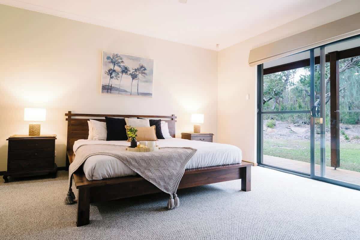 A bedroom with a large bed, two bedside tables, a painting on the wall, and a view through a sliding glass door.
