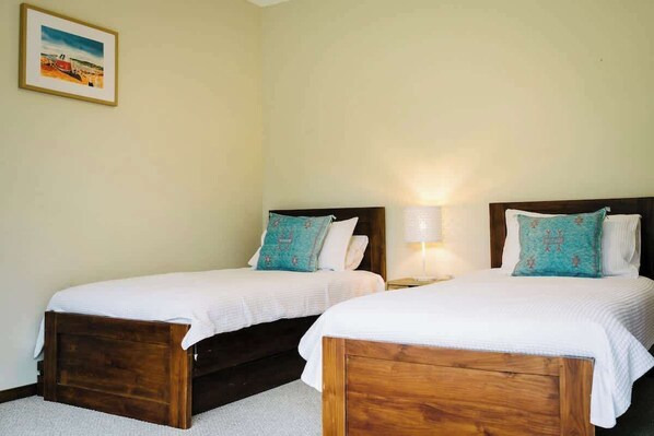 Une chambre avec deux lits en bois, chacun recouvert d’une courtepointe blanche et agrémenté d’un coussin turquoise. Un tableau encadré est accroché au mur, et une lampe de chevet se trouve sur le côté droit.