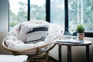 A wicker chair with a white cushion and a patterned throw blanket, a small round table with a plant, a pair of glasses, and a notebook.