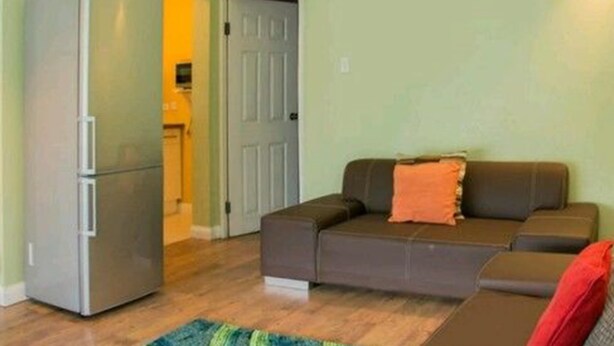 Ein kompakter Wohnraum mit einem grauen Kühlschrank, einem braunen Sofa mit orangefarbenem Kissen und Holzboden.