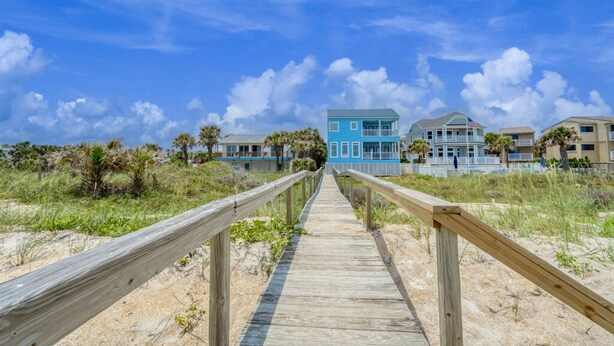 A wooden boardwalk leading to a beachfront property with multiple buildings, including a prominent blue house.
