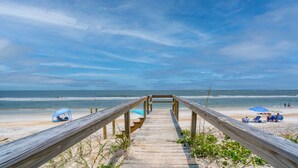 A wooden boardwalk leading to a beach with umbrellas and people.