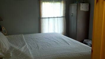 A bedroom with a bed, a window with curtains, a wardrobe, and a small table with a lamp.