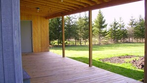 Eine Holzterrasse mit überdachtem Bereich, einer Tür und Blick auf einen grasbewachsenen Garten mit Bäumen.