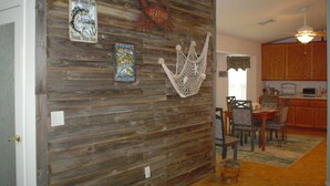 Un coin repas avec un mur en bois, un ventilateur de plafond et une cuisine en arrière-plan.