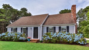 A single-story house with a brick chimney, white siding, and a front porch. - Unique Oyster Pond 3bd-2ba home in private setting close to downtown. (Chatham)